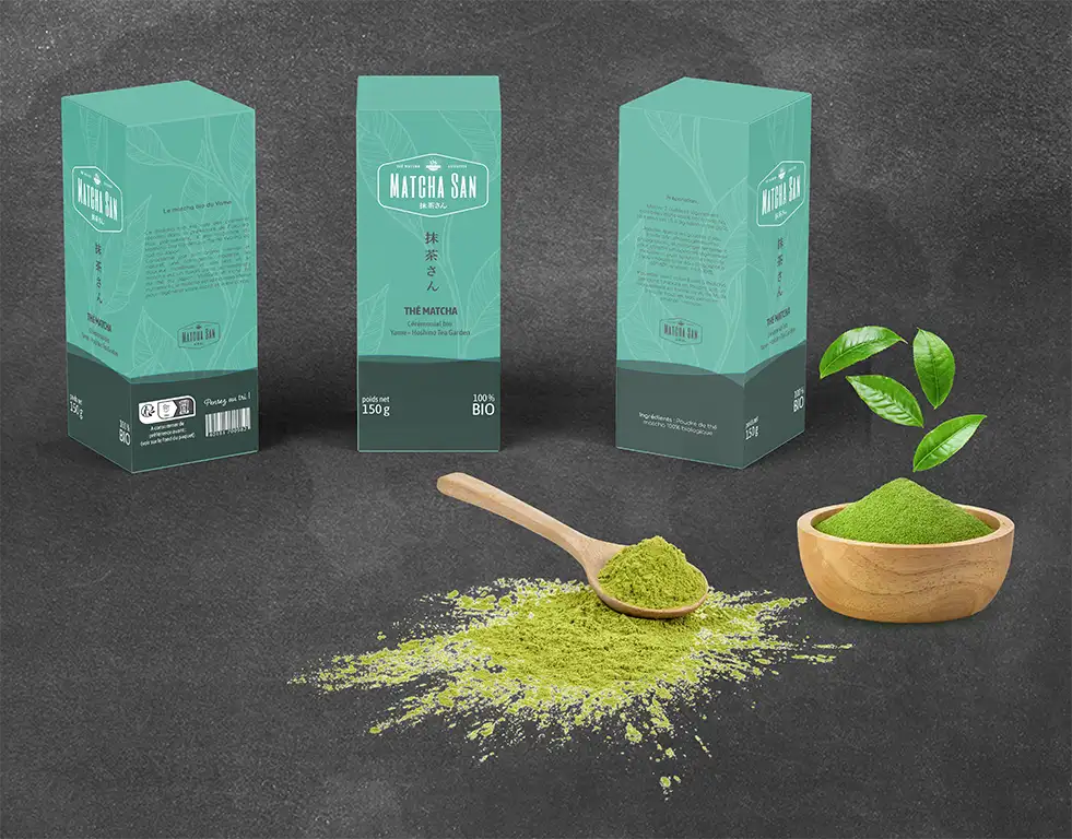 mock up de boites de thé Macha San. 3 boites sont posées sur une table noir. Il y a un tas de thé vert ainsi qu'une cuillère et un bol en bois.